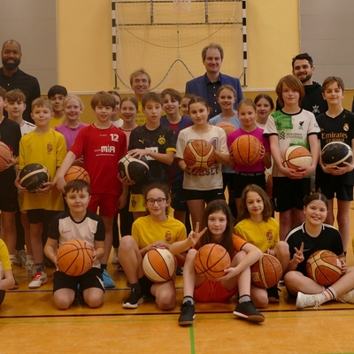 Sch&uuml;lergruppe mit Basketb&auml;llen posiert gemeinsam mit erwachsenen G&auml;sten in einer Sporthalle.