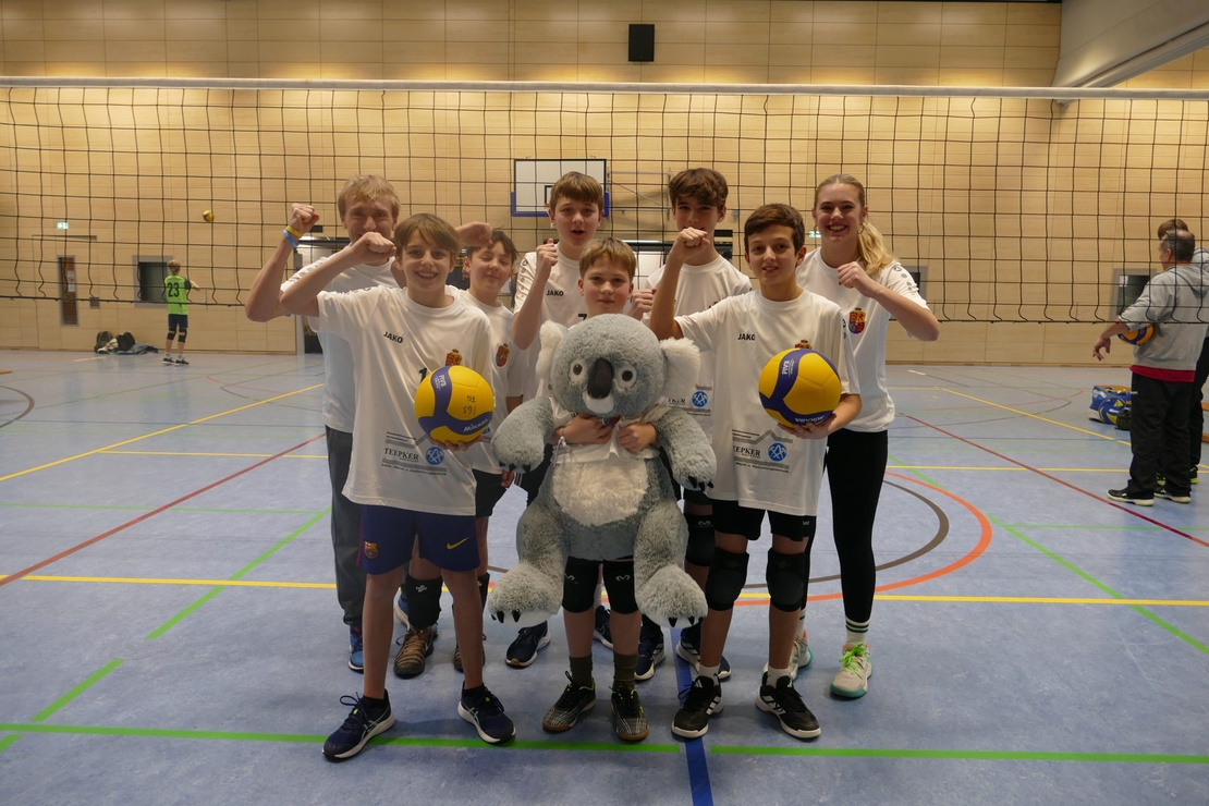 Jugendliche Volleyballmannschaft posiert mit zwei Volleyb&auml;llen und einem Koala-Maskottchen in einer Sporthalle.
