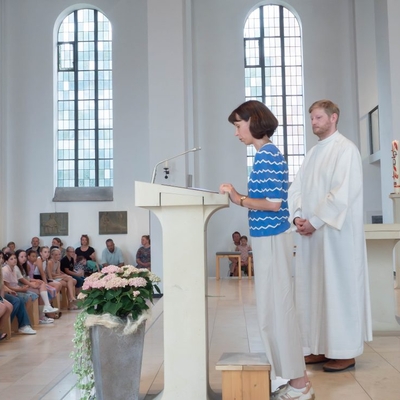 Eine Frau steht an einem Lesepult in der Kirche, daneben steht ein Priester.