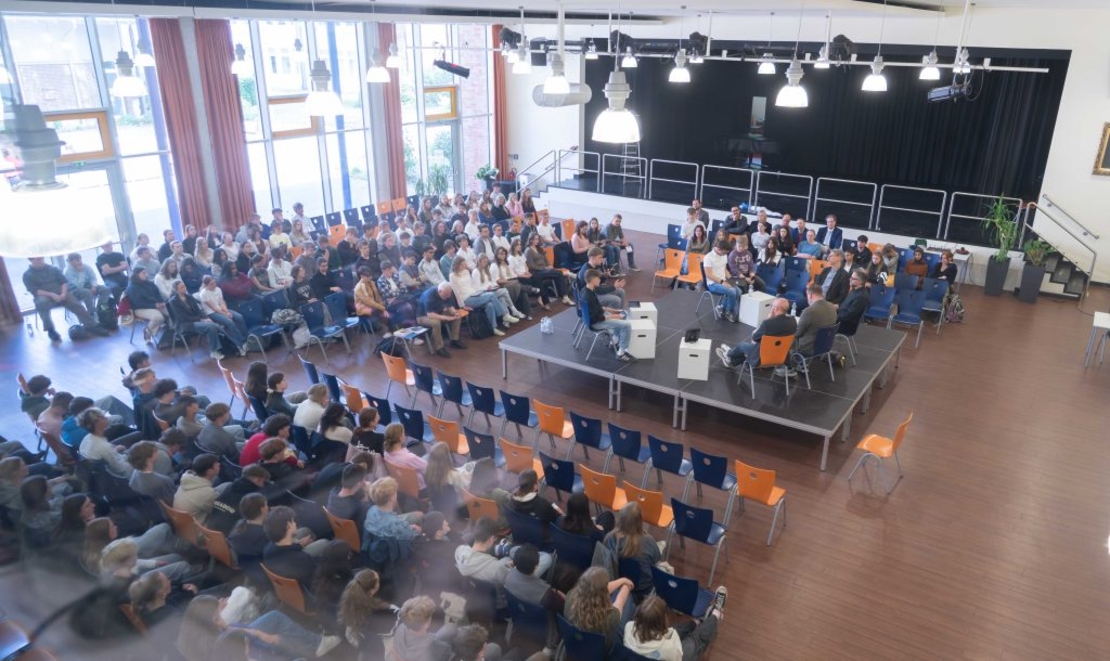 Blick von oben auf eine Podiumsdiskussion mit vielen Zuh&ouml;rerinnen und Zuh&ouml;rern in der Mensa.
