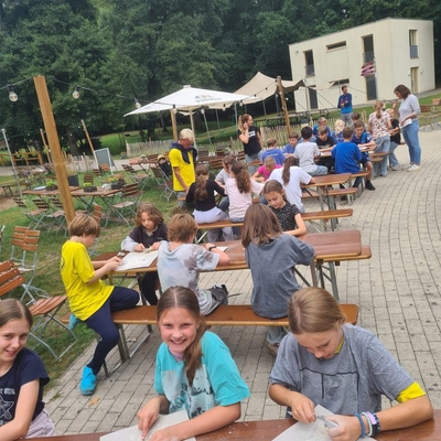 Kinder sitzen und stehen im Au&szlig;enbereich an Tischen und B&auml;nken.