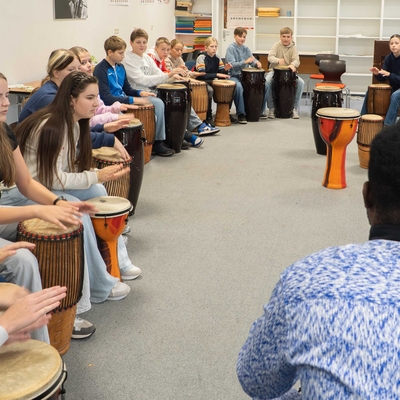 Sch&uuml;lerinnen und Sch&uuml;ler sitzen im Kreis und trommeln gemeinsam in einem Workshopraum.