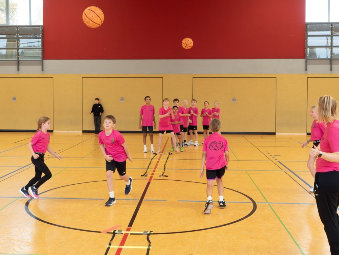 Kinder in pinken Shirts bewegen sich gemeinsam bei einer Sport&uuml;bung mit Basketb&auml;llen in einer Sporthalle.