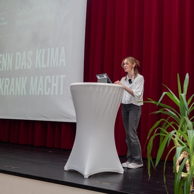 Eine Jugendliche h&auml;lt einen Vortrag auf einer B&uuml;hne vor gro&szlig;er Leinwand.