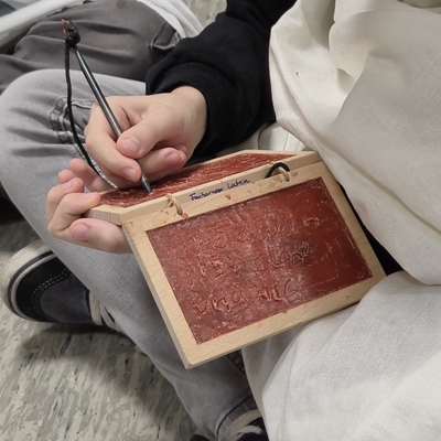 Eine Person schreibt mit einem Stift auf eine antik wirkende Schreibtafel.