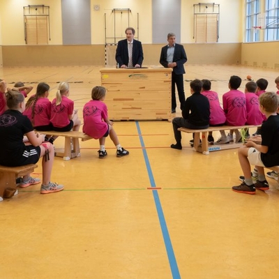 Kinder sitzen in einer Sporthalle und verfolgen einen Vortrag.
