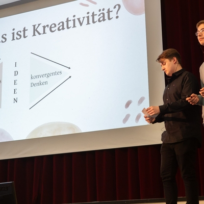 Zwei Jugendliche pr&auml;sentieren vor einer Leinwand mit der &Uuml;berschrift Was ist Kreativit&auml;t.