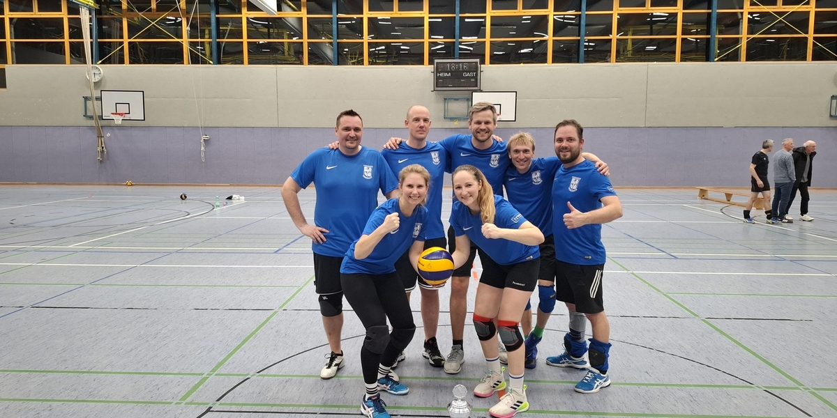 Lehrervolleyballteam in blauen Trikots posiert mit einem Volleyball in einer Sporthalle.