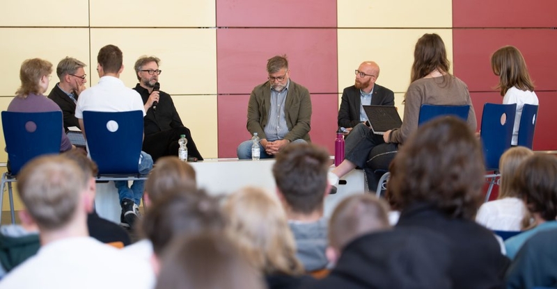 Podiumsgespr&auml;ch mit mehreren Erwachsenen vor einem Publikum in einem Veranstaltungsraum.