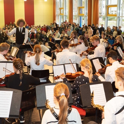 Sch&uuml;lerinnen und Sch&uuml;ler musizieren mit Streichinstrumenten vor Publikum in der Mensa.