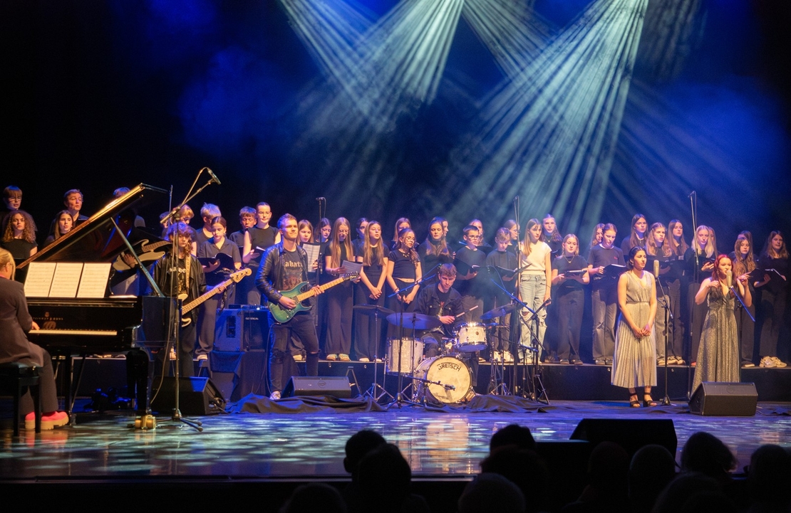 Gro&szlig;es Ensemble aus Chor, Band und Solistinnen steht bei einem Konzert auf einer beleuchteten B&uuml;hne.