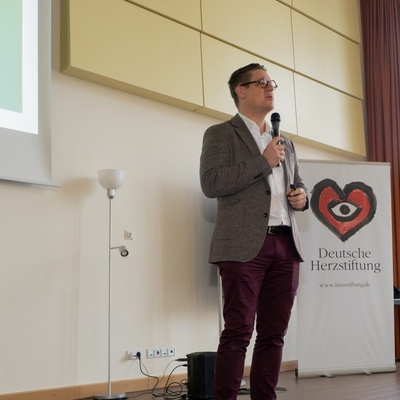 Ein Mann spricht mit Mikrofon neben einem Roll-up in einem Veranstaltungsraum.