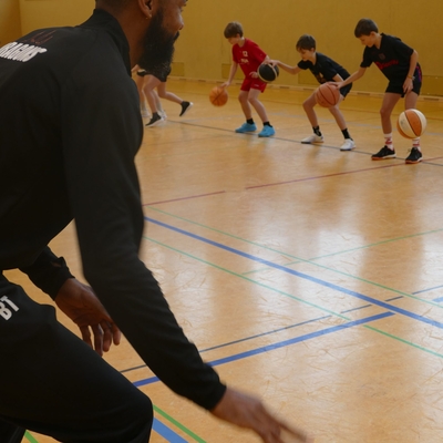 Trainer im Vordergrund beobachtet Jugendliche bei einer Basketball&uuml;bung in der Sporthalle.