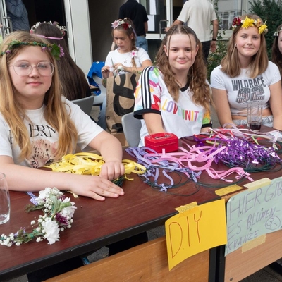 Sch&uuml;lerinnen sitzen an einem Stand mit Blumenkr&auml;nzen und Bastelmaterial.