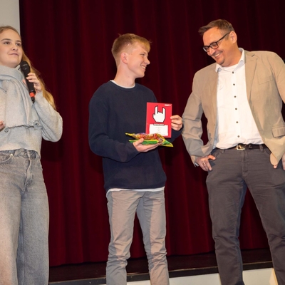 Zwei Jugendliche &uuml;berreichen ihrem Lehrer und Mentor auf der B&uuml;hne ein Geschenk.