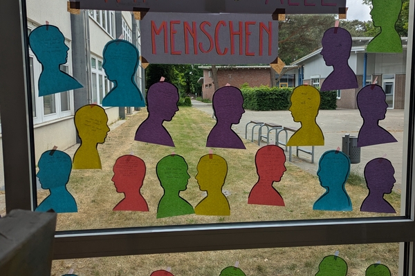 Bunte Papier-Silhouetten menschlicher K&ouml;pfe an einer Fensterscheibe, dar&uuml;ber die &Uuml;berschrift "Wir sind alle Menschen"