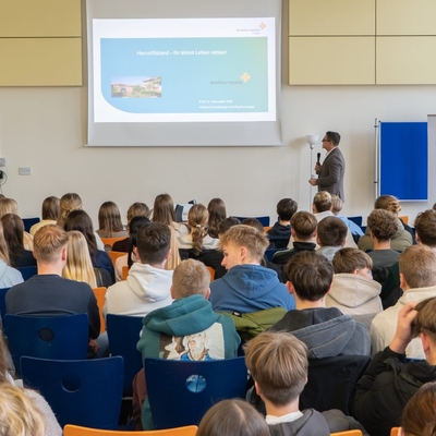Publikum verfolgt einen Vortrag mit Pr&auml;sentation in einem Veranstaltungsraum.