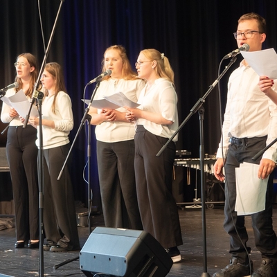 Jugendliche singen gemeinsam an Mikrofonen auf einer B&uuml;hne.
