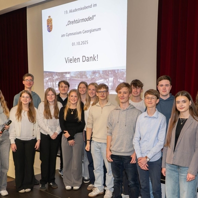 Gruppe von Jugendlichen posiert gemeinsam mit einem Lehrer auf einer B&uuml;hne vor einer Leinwand.