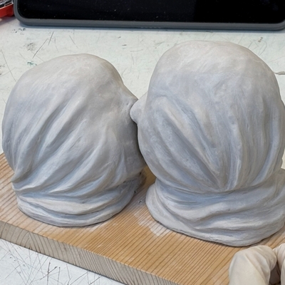 Zwei mit Stoff verh&uuml;llte Kopfskulpturen nebeneinander auf einem Tisch.