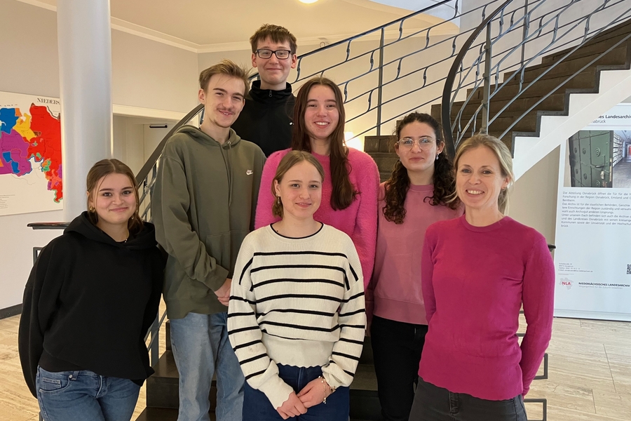 Gruppe von Jugendlichen und einer erwachsenen Begleitperson posiert in einem Archivgeb&auml;ude vor einer Treppe.