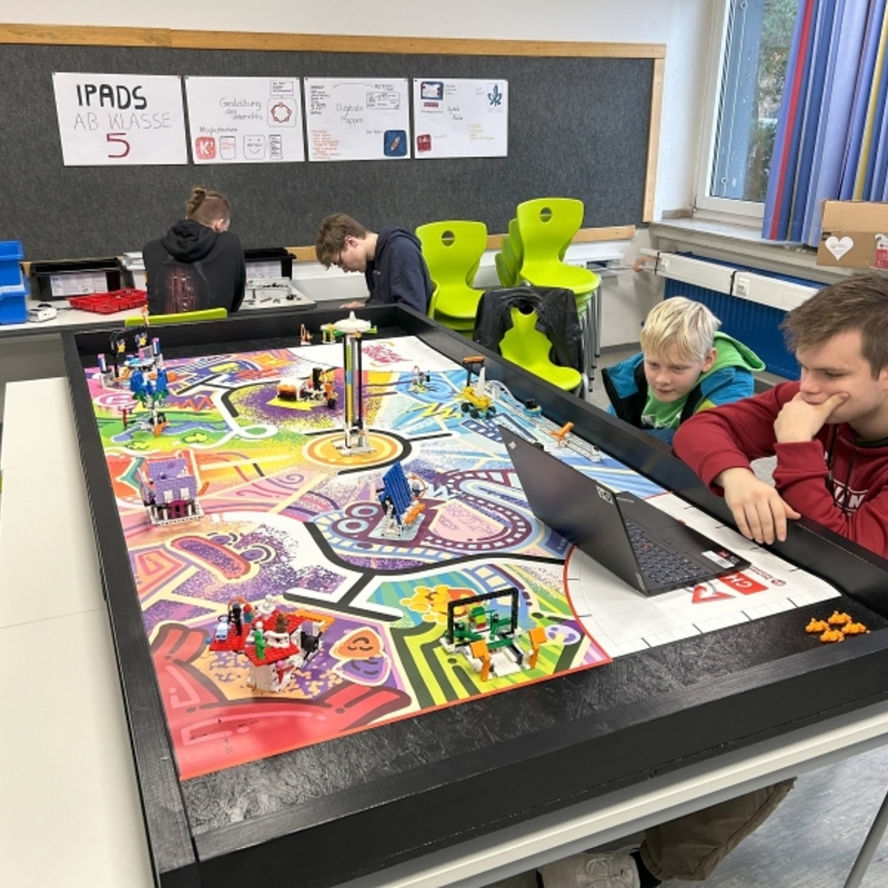 Sch&uuml;lerinnen und Sch&uuml;ler arbeiten mit einem gro&szlig;en Spielfeld und Robotikmaterial im Informatikunterricht.