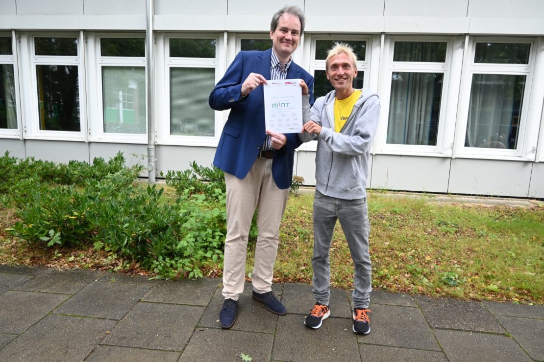 Zwei Lehrer posieren mit einem Zertifikat vorm Schulgeb&auml;ude.