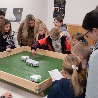 Kinder beobachten an einem Tisch einen kleinen Roboter auf einer gr&uuml;nen Fl&auml;che.