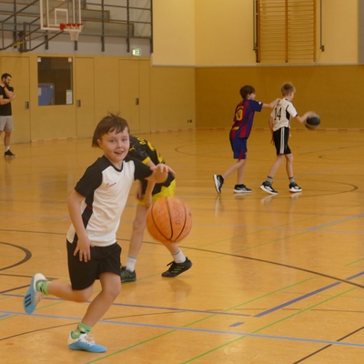 Sch&uuml;ler l&auml;uft mit einem Basketball durch die Sporthalle, im Hintergrund trainieren weitere Kinder.