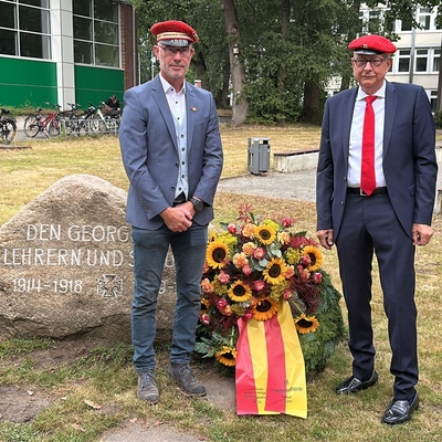 Zwei M&auml;nner mit roten M&uuml;tzen stehen neben einem Gedenkstein mit Blumenschmuck.