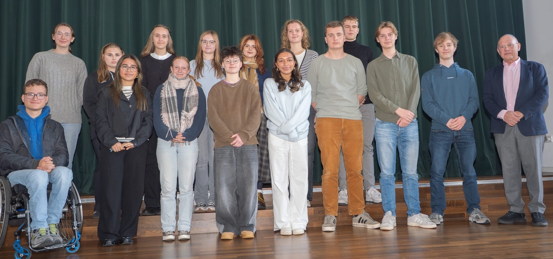 Sch&uuml;lerinnen und Sch&uuml;ler posieren mit einem erwachsenen Gast f&uuml;r ein Gruppenfoto auf einer B&uuml;hne.
