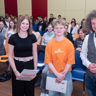 Zwei Jugendliche und ein Lehrer posieren f&uuml;r ein gemeinsames Foto in einem gut besuchten Veranstaltungsraum.