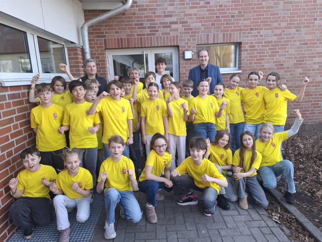 Sch&uuml;lergruppe in gelben T-Shirts posiert mit zwei erwachsenen Begleitpersonen vor einem Backsteingeb&auml;ude.
