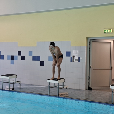 Person steht auf einem Startblock am Beckenrand eines Hallenbads.