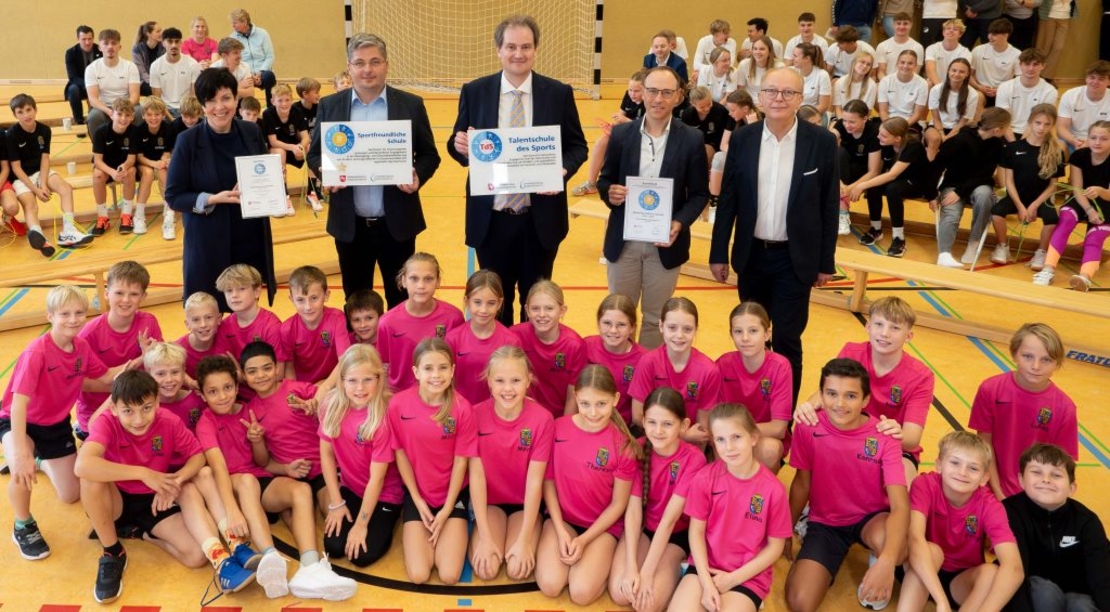 Kindergruppe in pinken Shirts posiert mit mehreren Erwachsenen, die Urkunden in einer Sporthalle halten.